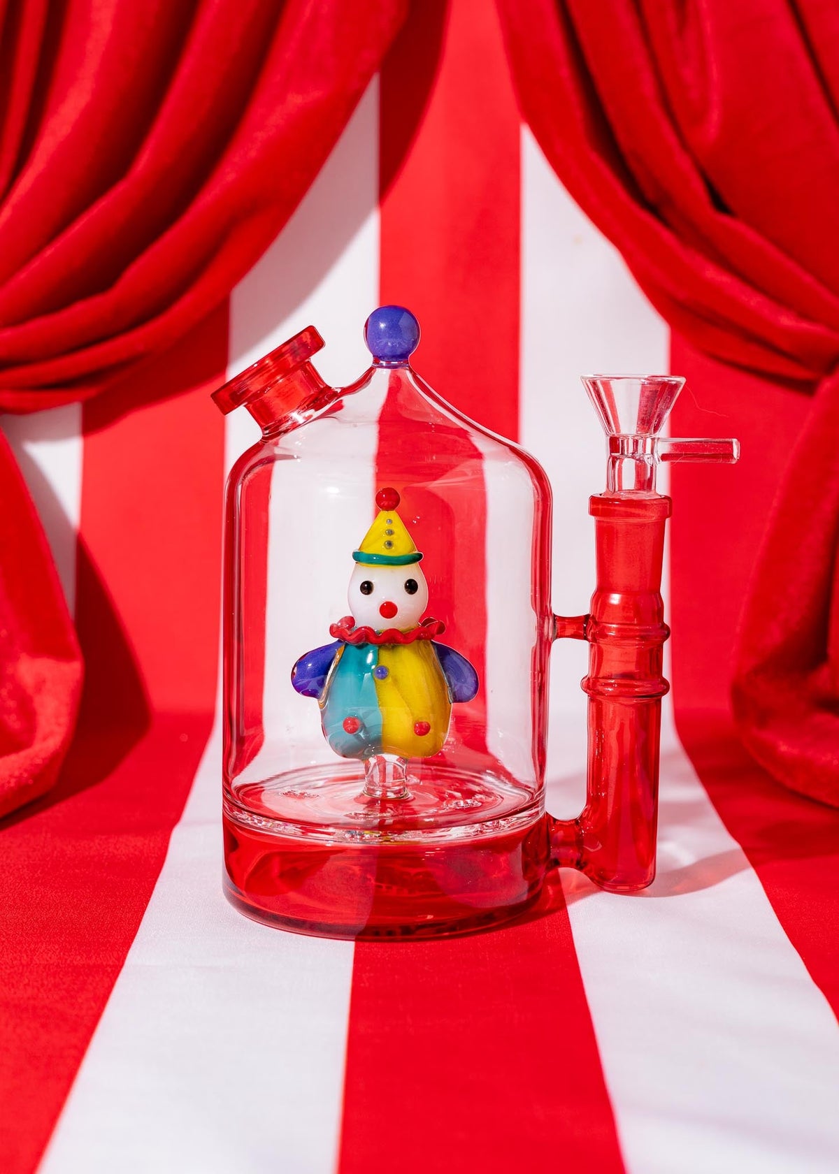 Colorful glass smoking device with a clown figure inside, against a red and white striped background.